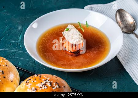 Suppe oder Consomme, dicke Fleischbrühe mit Karotten, Hähnchenknödel serviert auf einem weißen Teller auf dunkelgrünem Hintergrund mit Sesambrötchen, Serviette und Stockfoto