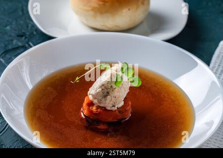 Suppe oder Consomme, dicke Fleischbrühe mit Karotten, Hähnchenknödel serviert auf einem weißen Teller auf dunkelgrünem Hintergrund mit Sesambrötchen, Serviette und Stockfoto