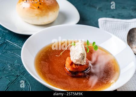 Suppe oder Consomme, dicke Fleischbrühe mit Karotten, Hähnchenknödel serviert auf einem weißen Teller auf dunkelgrünem Hintergrund mit Sesambrötchen, Serviette und Stockfoto