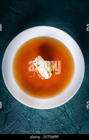 Suppe oder Consomme, dicke Fleischbrühe mit Karotten, Hähnchenknödel serviert auf einem weißen Teller auf dunkelgrünem Hintergrund mit Sesambrötchen, Serviette und Stockfoto