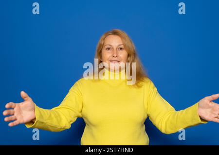 Weiße Frau in ihren 40ern, die einen gelben Pullover mit offenen Armen in einer einladenden Geste trug, isoliert auf blauem Atelierhintergrund. Stockfoto
