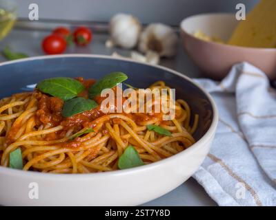Balanesi-Pasta mit Basilikumblättern und Tomatensauce in einer Schüssel serviert, mit Zutaten wie Tomaten, Knoblauch und Käse im Hintergrund Stockfoto