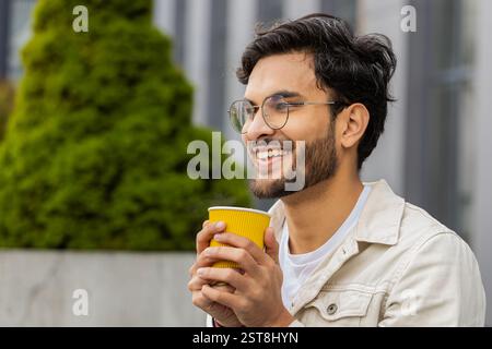 Glücklicher junger indischer Mann, der morgens Kaffee, heißes Getränk genießt und draußen lächelt. Entspannen, eine Pause einlegen. Ein arabischer Hindu-Typ in der Innenstadt, trinkt Kaffee zum Mitnehmen. Stadtleben draußen Stockfoto