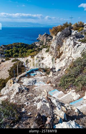 Ein zerklüfteter Steinweg mit blau gemalten Stufen führt einen felsigen Hügel hinunter, mit Blick auf den korinthischen Golf und den Melagavi Leuchtturm, mit einem Kreuz auf dem Stockfoto