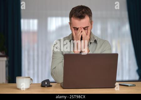Ein müder Mann arbeitete Überstunden, saß am Schreibtisch vor einem Laptop und reibte sich die Augen mit den Händen. Stockfoto