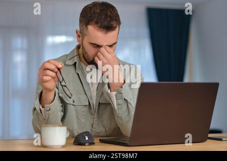 Müder Mann arbeitet Überstunden, sitzt am Schreibtisch vor einem Laptop, hält die Brille und reibt sich die Augen mit der Hand. Stockfoto