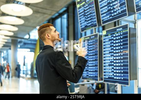 Geschäftsmann, der Smartphone hält und auf die Fahrplananzeige zeigt, Fluginformationen im Flughafenterminal überprüft Stockfoto