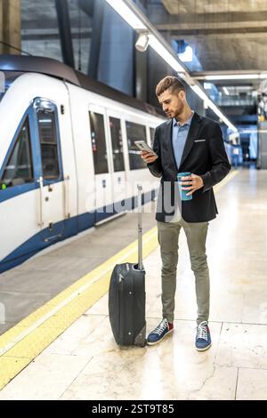 Geschäftsmann steht auf U-Bahn-Bahnsteig, benutzt Smartphone, hält Kaffee und wartet mit seinem Wagen auf den Zug Stockfoto