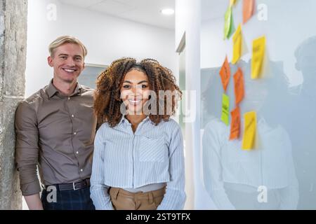 Zwei lächelnde Kollegen stehen in einem Coworking-Büro in der Nähe eines Whiteboards mit bunten Haftnotizen Stockfoto