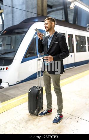 Junger Geschäftsmann, der auf die U-Bahn wartet, Smartphone benutzt und eine wiederverwendbare Kaffeetasse hält, mit Koffer neben ihm Stockfoto