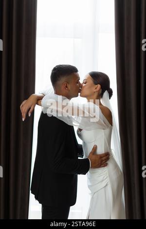 Romantisches Porträt des Bräutigams, der Braut auf die Stirn küsst, während er sich am Hochzeitstag in der Nähe des Fensters umschließt Stockfoto