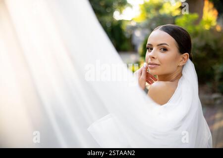 Schöne Braut posiert anmutig mit einem fließenden weißen Schleier, strahlt Freude und Eleganz an ihrem Hochzeitstag in einer ruhigen Parklandschaft aus Stockfoto