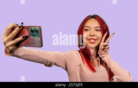 Lächelnder Influencer zeigt ein Friedenszeichen, während er ein Selfie mit Smartphone auf violettem Hintergrund macht Stockfoto