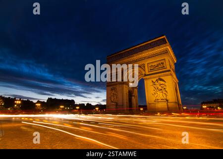 Frankreich, Paris. Der Arc de Triomphe steht vor einem dramatischen Dämmerhimmel, mit goldenen Lichtspuren vorbeifahrender Fahrzeuge, die die Lichter unterstreichen Stockfoto