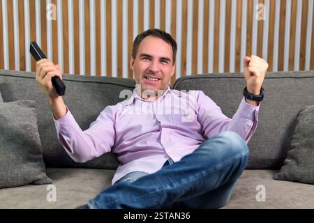 Positiver, fröhlicher Mann, der zu Hause sitzt und Fußballspiele ansieht, entspannen Sie sich drinnen. Der glückliche Kerl auf der Couch schaut mit der Fernbedienung in den Händen fern. Stockfoto