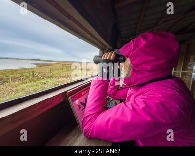 Eine Frau in einer leuchtend rosa Jacke, die durch ein Fernglas in einem Vogelhäuschen im Naturschutzgebiet Blacktoft Sands schaut. Goole. UK Stockfoto