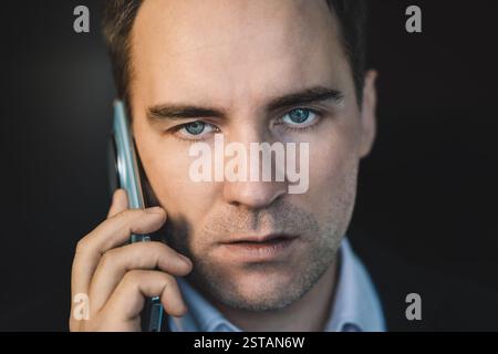 Ein hübscher Geschäftsmann mit einem verächtlichen Blick spricht nachts auf dem Handy vor dunklem Hintergrund. Nahporträt eines kaukasischen Mannes mit grauem stubb Stockfoto