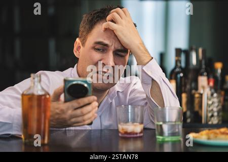Ein junger Mann in einem Hemd sitzt in einer Bar, trinkt Whiskey und liest eine Nachricht auf einem Smartphone mit Traurigkeit im Gesicht. Das Konzept des persönlichen PR Stockfoto