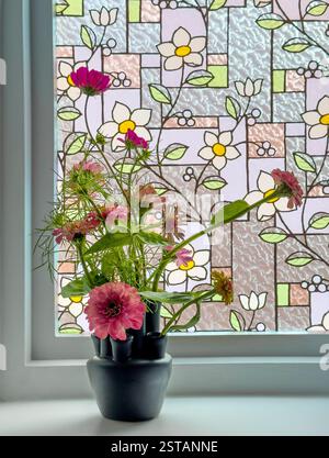 Schwarze Vase mit mehreren Ausläufen und rosa Zinnien und Kosmos auf einer weißen Fensterbank, die vor einem Glasfenster mit Blumenmuster steht Stockfoto