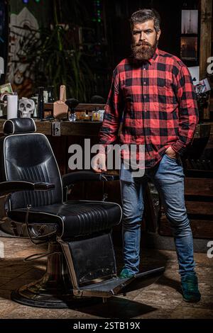 Mann Barbier im Hemd, der neben dem Stuhl im Friseursalon steht. Ein bärtiger Mann steht im Friseur. Friseur steht auf einem Barbierstuhl an seinem Arbeitsplatz Stockfoto