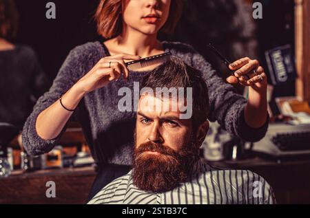 Frau Friseur mit einer Schere unterschneiden. Friseurmädchen, der den bärtigen Mann im Friseurladen frisiert, Pflege, männliche Schönheit Stockfoto