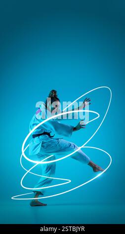 Weibliche Kampfkünstlerin in weiß, die Verteidigungsstellung ausübt, neonblaue Lichtspuren, die sich um Präzision und Balance vor blauem Hintergrund bewegen. Stockfoto