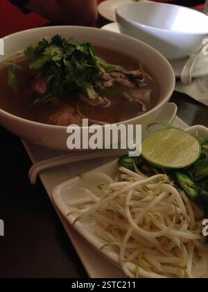 Schüssel Pho, Teller Bohnensprossen, Limettenscheibe auf dem Tisch. Pho ist vietnamesische Nudelsuppe mit reichhaltiger Rinderbrühe und dünnen Reisnudeln. Zufriedenstellendes Mittagessen Stockfoto