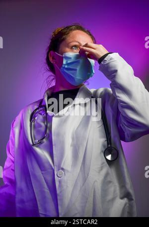 Eine Ärztin trägt eine chirurgische Maske und einen weißen Mantel mit Stethoskop und berührt ihr Gesicht unter purpurrotem Licht Stockfoto