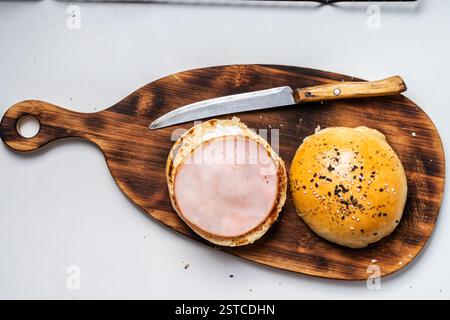 Sesambrötchen geschnitten, um Schinken und Käse Sandwich auf hölzernem Schneidebrett mit Messer auf grauem Hintergrund zu machen. Hochwertige Fotos Stockfoto