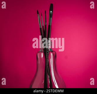 Farbenfrohe Ausstellung von Künstlerbürsten in einer Glasflasche vor leuchtendem rosa Hintergrund Stockfoto