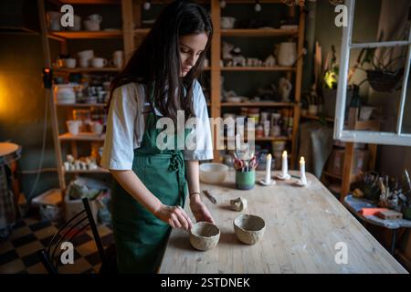 Herstellung von handwerklicher Keramik. Arbeitstisch im Keramikstudio mit Frau, die nassen Tonbecher formt Stockfoto