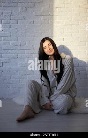 Eine junge Frau mit langen dunklen Haaren sitzt bequem auf dem Boden und trägt einen hellen Pyjama. Sanftes natürliches Sonnenlicht erhellt ihren ruhigen Ausdruck Stockfoto