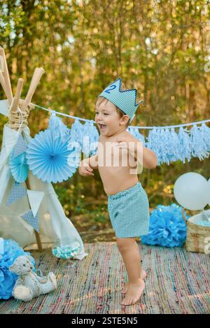 Ein fröhlicher Kleinkind in einer gemusterten Krone und Shorts feiert seinen zweiten Geburtstag im Freien mit einem dekorierten Zelt, blauen Rädern und Ballons i Stockfoto