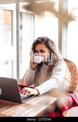 Junge Freiberuflerin genießt Tee und arbeitet am Netbook, während sie zu Hause am Tisch sitzt, während sie in einer modernen Wohnung unterwegs ist Stockfoto