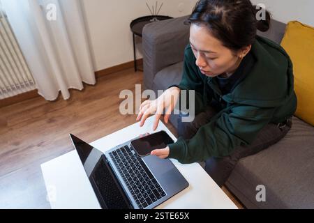 Eine fokussierte Frau arbeitet von zu Hause aus, verwendet einen Laptop, während sie ein Smartphone in der Hand hält, und stellt Multitasking und digitale Konnektivität vor Stockfoto