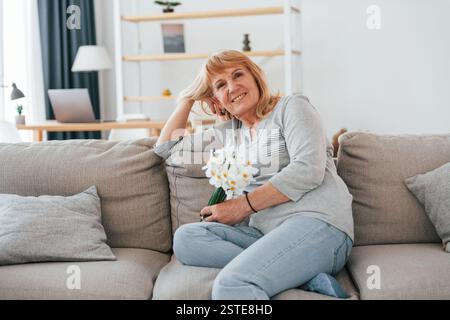 Auf einem Sofa sitzen, Blumen in den Händen. Eine Seniorin mit roten Haaren ist allein zu Hause. Stockfoto