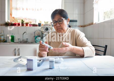 Eine ältere Frau sitzt an einem Küchentisch und konzentriert sich, während sie mit einem Glukometer ihren Blutzucker kontrolliert, umgeben von medizinischen Vorräten und einem Heim Stockfoto