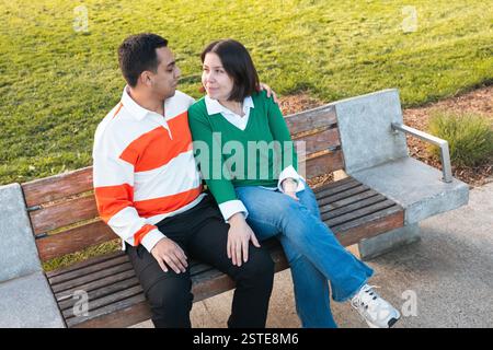 Ein junges lateinisches Paar sitzt eng auf einer Parkbank und teilt einen Moment der Zuneigung mit einem Lächeln und einem sanften Blick in einer ruhigen Umgebung im Freien Stockfoto