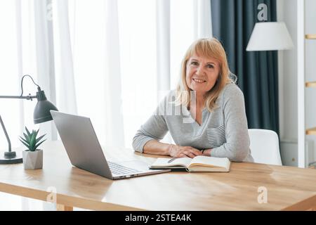 Mit Laptop am Tisch sitzen. Eine Seniorin mit roten Haaren ist allein zu Hause. Stockfoto