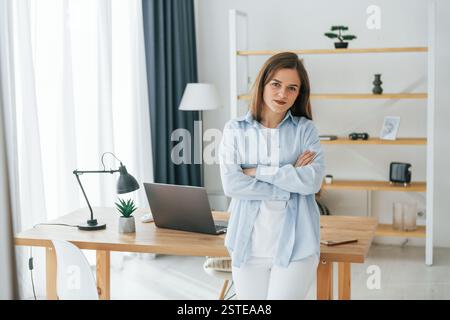 Stehend mit gekreuzten Armen. Die junge schöne Frau ist allein zu Hause im Wohnzimmer. Stockfoto