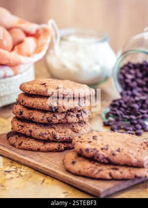Ein rustikaler Tisch hält einen Stapel frisch gebackener Schokoladenkekse neben Zutaten wie Eiern, Mehl und Schokoladenchips, die die Wärme o verkörpern Stockfoto