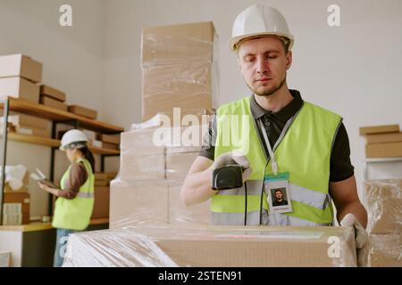 Arbeiter in einem Lager, die Kisten auspacken und Sicherheitswesten und Helme tragen, während sie Barcode-Scanner verwenden, Regale mit Kisten und Industrieausrüstung umgeben Stockfoto