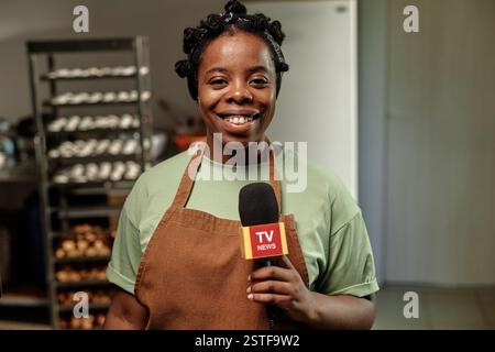 Porträt einer lächelnden afroamerikanischen Interviewpartnerin mit Mikrofon, die mit Journalisten über die Arbeit in einer kleinen Bäckerei spricht, die in der Küche steht, im Kopierraum Stockfoto