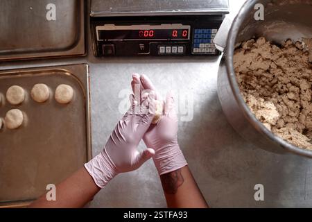 Bild von oben von unerkennbaren schwarzen Frauen Hände in Nitril Einweghandschuhen rollendes Stück rohen Teigs, der runde Weizenbrötchen auf der Arbeitsstation in der Bäckereiküche macht, Kopierraum Stockfoto