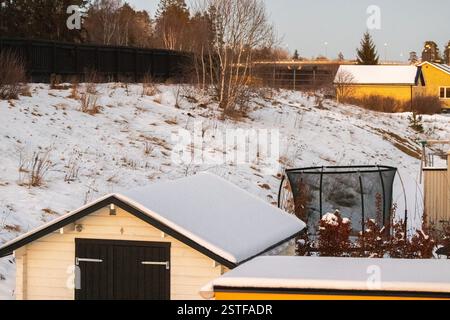Kleiner Holzschuppen mit schneebedecktem Dach in der Nähe von Hügeln mit Bäumen und getrocknetem Gras am kalten Winterabend. Schweden. Stockfoto