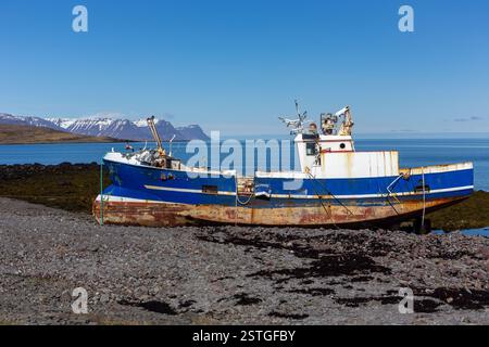 Altes, rostiges Stahlfischboot, das am Ufer der Westfjorde, Island, mit schneebedeckten Bergen im Hintergrund liegt. Stockfoto