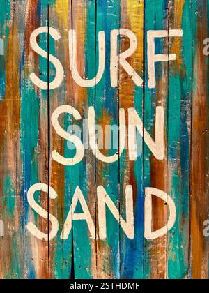 Lebendige und rustikale Kunstwerke mit den Worten „Surf Sun Sand“, die in weiß auf einem strukturierten Holzbrett gemalt sind. Die Türkis-, Gold- und Brauntöne erinnern an Stockfoto