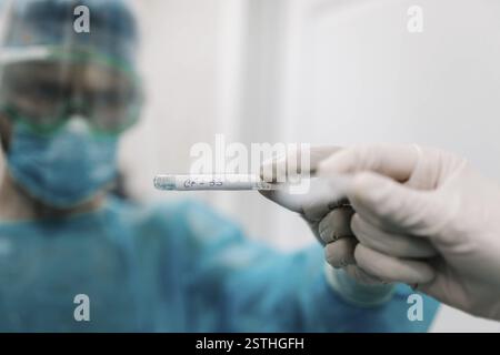 Eine Hand mit Handschuhen, die ein Reagenzglas in einer Laborumgebung hält, mit einer maskierten Person im Blick Stockfoto