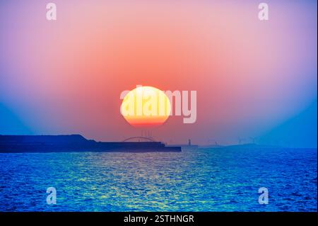 Ein atemberaubender Sonnenuntergang über dem Tamsui-Meer mit leuchtenden Farben, die sich am Himmel vermischen. Silhouetten von Windturbinen und eine Brücke verleihen der Szene Tiefe. Tamsu Stockfoto