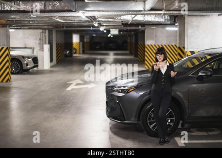 Selbstbewusste asiatische Frau in den 30ern, die auf schwach beleuchteten Parkplätzen eine Sprachnachricht schickte. Professionelle Kleidung ist ein geschäftliches Umfeld, da sie sich mit dem Telefon in der Hand gegen ein modernes Fahrzeug lehnt. Stockfoto